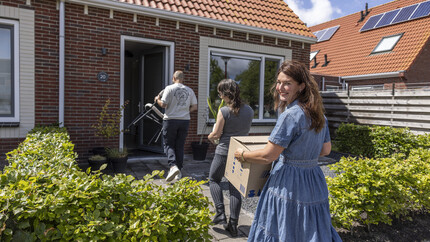 Mevrouw die een verhuisdoos naar de voordeur tilt en glimlachend achterom kijkt